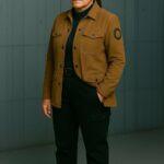 Une femme debout, vêtue d’une veste brune et d’un pantalon noir, les mains dans les poches, posant avec assurance dans un décor sobre et moderne | A woman standing, wearing a brown jacket and black pants, hands in her pockets, posing confidently in a minimalist modern setting